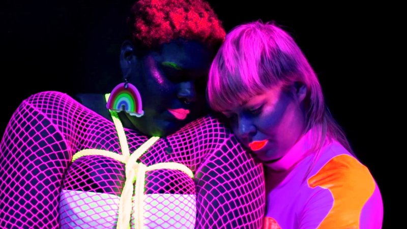 two women bathed in bright neon standing close, one is wearing rope bondage.