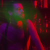 Still image from the short film, Nightender showing a shirtless man, bathed in red light, in a bar with rows of bottles behind him. 