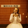 Still from the short film, Sologamy, of a woman in a wedding dress applying make-up to her face while waiting for a train. Wedding is spelled out on the metro station tiles behind her.g